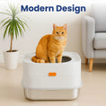 Load image into Gallery viewer, A modern top entry cat litter box with an orange button, featuring an orange tabby cat sitting on top, accompanied by a potted plant and a gray armchair.