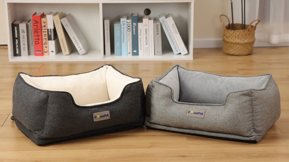 Two empty PawsPik heated dog beds on a wooden floor, one dark grey and one light grey, with a bookshelf in the background.