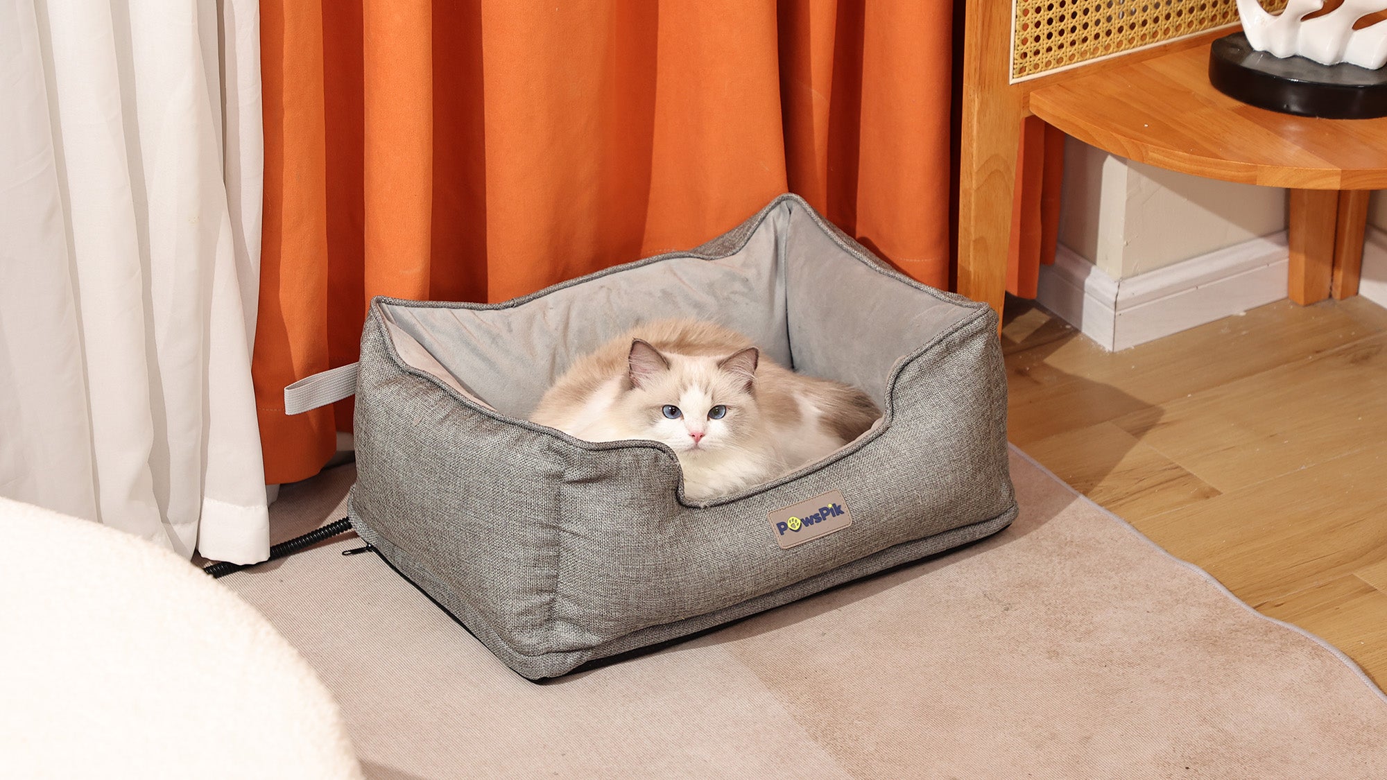 A fluffy white and beige cat resting in an ash-grey heated pet bed beside orange curtains.