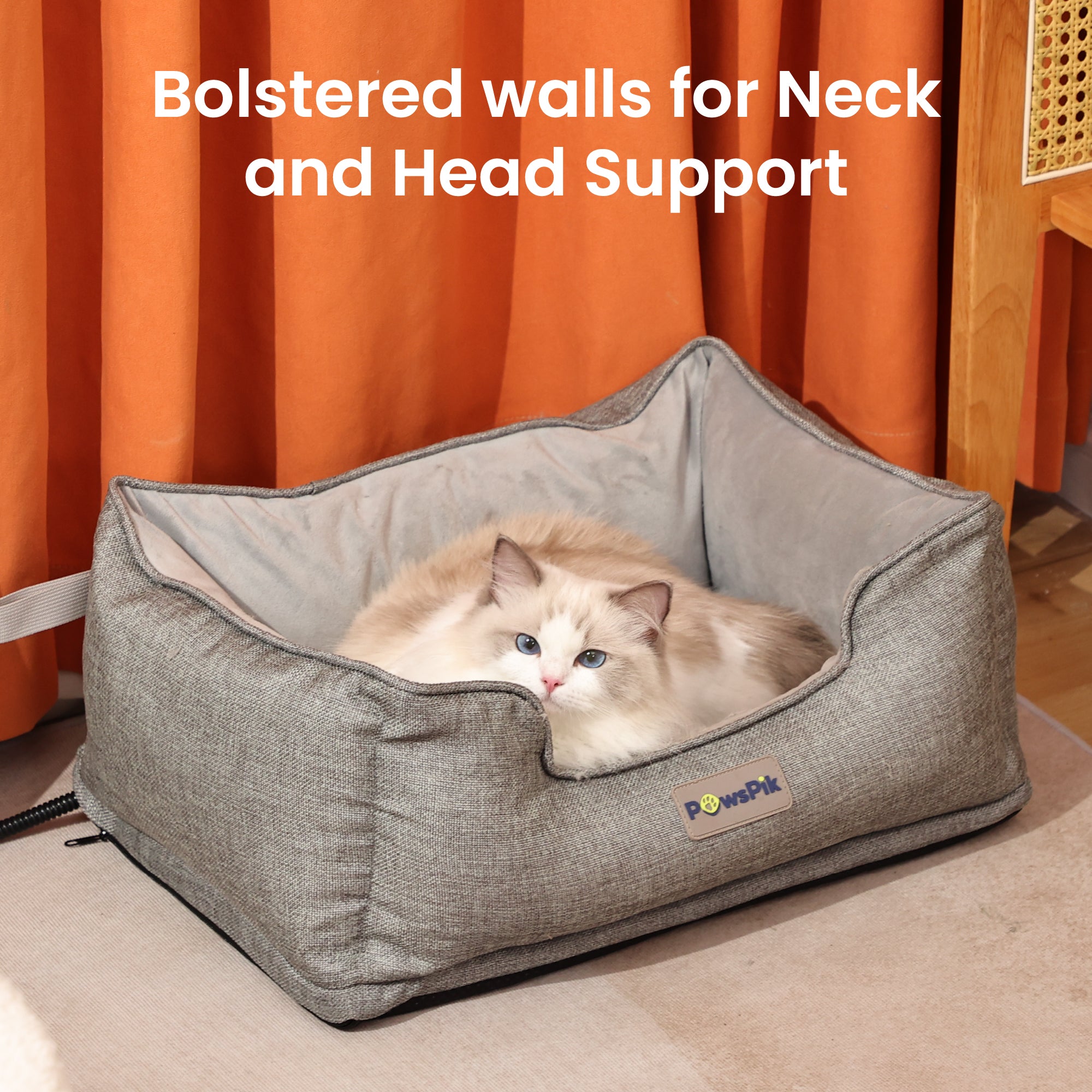 A fluffy white cat resting in an ash-grey heated cat bed by PawsPik against an orange curtain background with text “Head and Neck Support”.