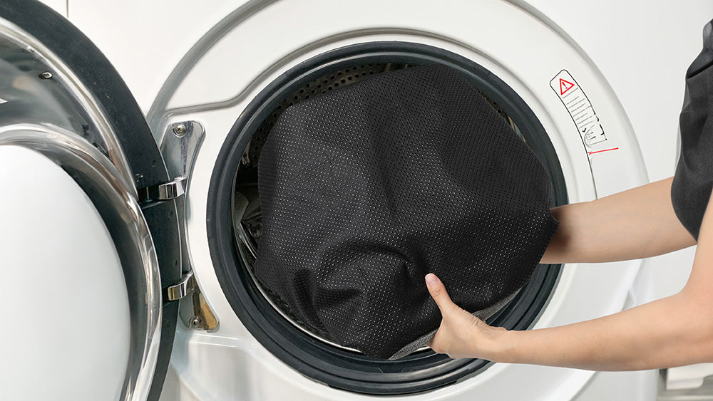 Person loading a black mesh heated dog bed cover into a washing machine.