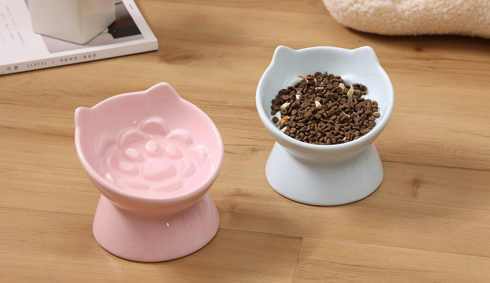 Two cute cat-shaped cat food bowls on a wooden surface: a pink bowl is empty, while a blue bowl is filled with pet food.