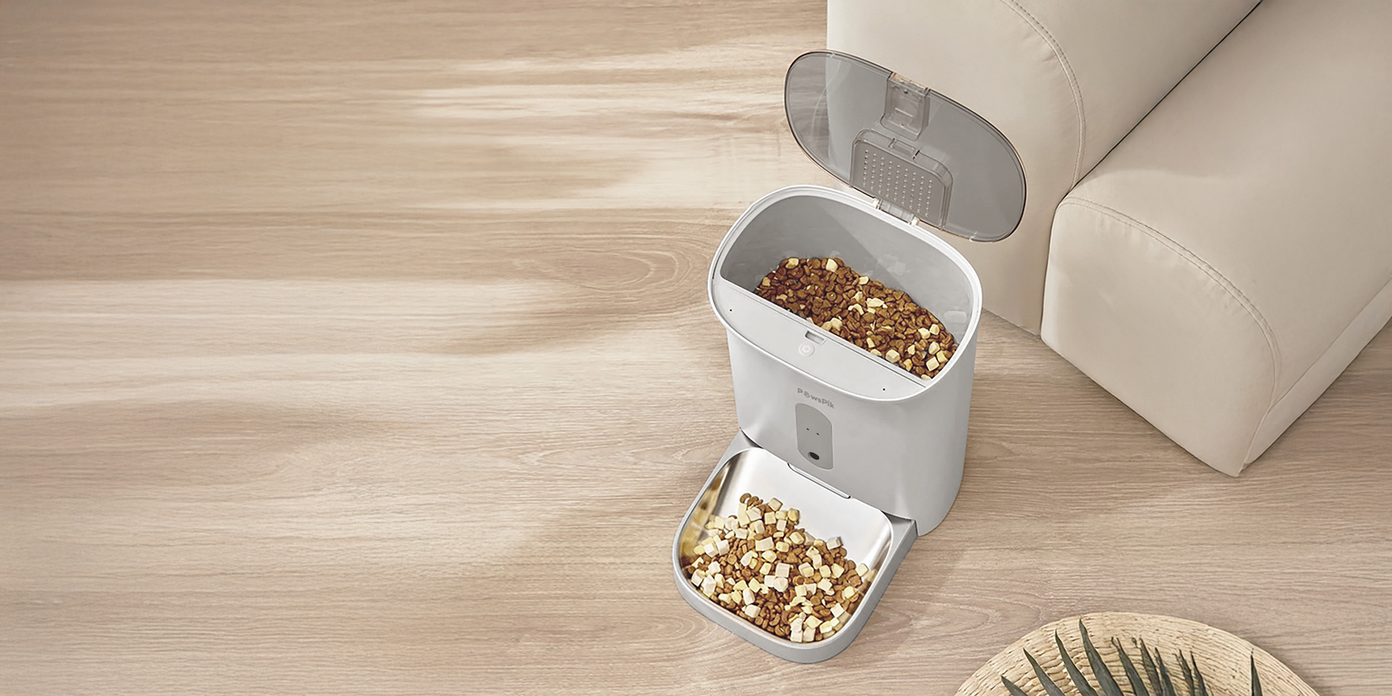 Best Automatic Cat Feeder filled with kibble is on a light wooden floor next to a cream sofa. Some leaves are partially visible in the corner.