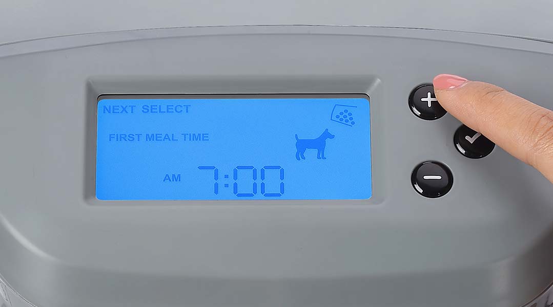 Finger pressing a button on the Pawspik Automatic pet feeder with a digital clock reading 'FIRST MEAL TIME 7:00 AM.